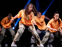 Performance énergique de danse urbaine : une danseuse aux tresses dynamiques, vêtue d’un t-shirt orange et d’un pantalon cargo, est capturée en plein mouvement sur scène.