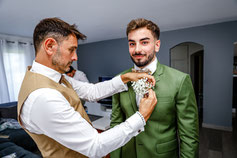 Un homme ajuste la boutonnière d'un marié souriant, vêtu d'un costume vert, dans une pièce au décor moderne.
