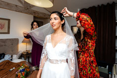 La mariée en robe blanche se prépare avec l'aide de ses proches, ajustant son voile dans une ambiance chaleureuse et émotive.
