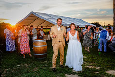 Les mariés avancent main dans la main, rayonnants de bonheur, sous le regard joyeux de leurs invités réunis pour célébrer cette belle soirée en plein air.