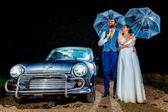 Photographe mariage dans le Tarn-et-Garonne couple de mariés sous la pluie avec parapluies devant une voiture ancienne de nuit