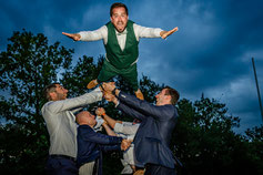 Un marié en costume vert projeté en l'air par ses amis, les bras grands ouverts et un sourire radieux, sous un ciel crépusculaire. Une explosion de joie et de complicité !