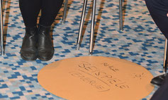 die Ergebnisse eines World Cafes sind auf eine runde hellbraune Pappe von ca. 50 cm Durchmesser geschrieben: in groß das Wort Beispiele, kleiner in Klammern Energie, eine gemalte Sonne, daneben die Schuhe von zwei Teilnehmern auf buntem Teppich