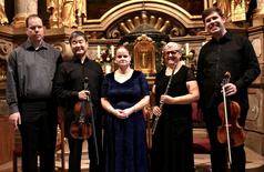 Zu sehen ist das Sonnenorchester, versammelt in der historischen Schlosskirche Mirabell. Das Ensemble steht inmitten des barocken Kirchenschiffs und bereitet sich auf seinen Auftritt vor.