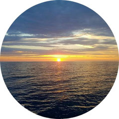 Sonnenaufgang über dem ruhigen Meer mit goldenen Lichtstrahlen am Horizont unter leicht bewölktem Himmel.