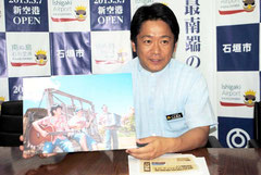 ビギンの写真を掲げ、新空港開港のPRソング制作を発表する中山市長=12日午後、市役所