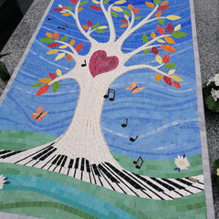Une  grande mosaïque sur une tombale représentant un arbre de vie  blanc dont les racines forment un clavier de piano noir et blanc, sur un fond de ciel bleu avec des notes de musique., 