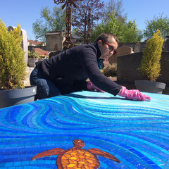 Une artiste mosaïste en plein travail, penchée sur la dalle funéraire pour nettoyer et ajuster les joints de la mosaïque bleue au cimetiere