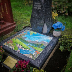 "Mosaïque funéraire paysagère sur monument en granit labrador bleu, décor de montagne avec rivière et fleurs sauvages, réalisée en émaux de Briare."