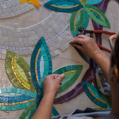 Gros plan sur les mains de l'artiste mosaiste en train d'assembler minutieusement des tesselles de mosaïque aux reflets verts et bleus pour former des feuilles.