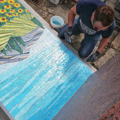 artiste mosaïste en plein travail de pose  d'une mosaïque représentant un ciel bleu et des tournesols à la Vangogh sur une pierre tombale.