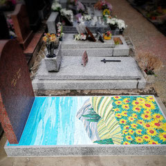 Vue d'ensemble d'un monument funéraire en Tarn gris orné d'une  mosaïque colorée représentant un paysage lumineux avec un champ de tournesols, installée en hommage à un artiste peintre défunt.