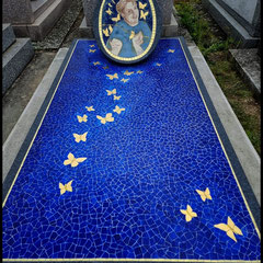 "Vue d'ensemble d'un monument funéraire en mosaïque bleu azur parsemé de papillons dorés s'élevant vers un médaillon central."