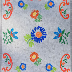 Gros plan sur le détail de la mosaïque funéraire mettant en valeur une fleur bleue centrale entourée de motifs de feuillages verts et de fleurs rouges sur fond gris.