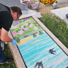Artiste mosaïste posant minutieusement le décor paysager aux hirondelles sur la pierre tombale en granit rose.