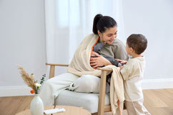 Mutter im Stuhl sitzend. Kind mit Baby im Stillschal von LÄSSIG. Kleinkind schaut zu.