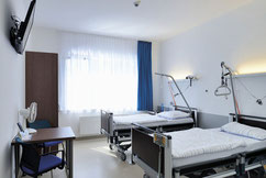 Zimmer im Medias Klinikum mit zwei Betten, einem Tisch, einem Stuhl und einem Fernseher an der Wand. Natürliches Licht durch das Fenster sorgt für eine helle, freundliche Atmosphäre.