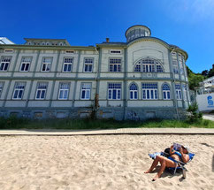 Woman in blue bikini on blue reclining chair in front of historic wooden bathing pavilion on beach in Jurmala, Latvia