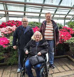 Two men standing in front of an elderly woman in a wheelchair in front of colourful azaleas in Riga's Botanical Gardens
