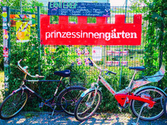 auf dem Foto zu sehen: zwei Fahrräder vor einem Bauzaun, dahinter ist alles sehr grün, oben drüber steht auf rotem Untergrund: Prinzessinnengärten