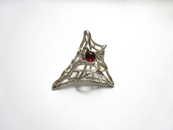 Une très grande bague ajourée au design semblable à une toile d'araignée avec en son centre un petit grenat rouge taillé en cabochon est exposée sur fond blanc.