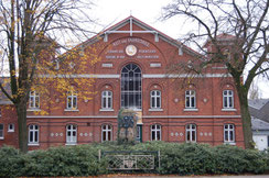 Historisches, rotes Backsteingebäude des Holsteiner Verbandes mit großen Fenstern und einem Wappen über dem Eingang, umgeben von Bäumen und Sträuchern.