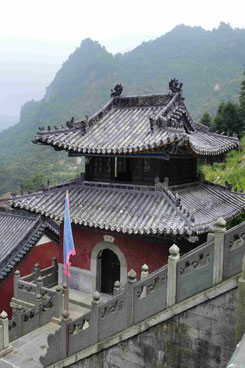 Sie sehen den Turm einer Tempelanlage in den heiligen Wudangbergen von meiner Chinastudienreise 2017. Der Turm ist dunkelrot mit zwei schönen Gibeldächern in unterschiedlichen Höhen. Im Vordergrund sieht man eine Fahne, im Hintergrund die grünen Berge.
