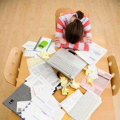 Jeune femme assise, la tête posée sur une table remplie de papiers, de dossiers et de post-it. La femme est brune et porte un pull rayé blanc et rouge.