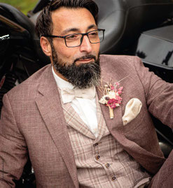 Man mit schwarzen Haaren und dunklem Vollbart unnd Brille sitzt entspannt in einem Auto. Er trägt einen Rostroten Anzug mit passender karierter Weste, Einstecktuch und anstecksträußchen. 