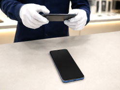 店舗カウンターに置かれたスマートフォンを、スタッフが別のスマホで撮影し、画面や本体の状態を確認している様子。スマホ買取や無料査定、店舗持ち込み査定に関連するイメージ。