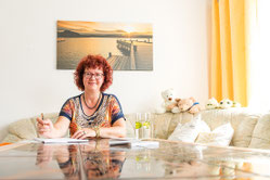 Ines Illing sitzt am Tisch, an der Wand ein Bild mit einem Steg am See
