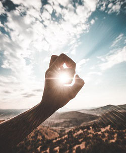 A hand, fingers curling, held out in front of the sun. The light of the sun shines through. The sky is blue with white clouds, and mountainous terrain stretches out ahead of the viewer.