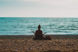 Une femme méditant assise en tailleur sur le sable face à la mer et au loin la ligne d'horizon