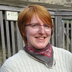 Ein Bild von mir bis zu den Schultern. Ich habe rötliche, kurze Haare, trage einen blass khakifarbenen Pullover und ein rot gemustertes Halstuch. Im Hintergrund sieht man hölzerne Gatter.