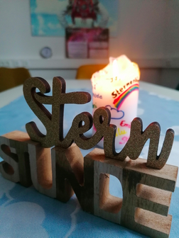 Auf einem Tisch steht im Vordergrund ein Dekoelement aus Holz das "Sternstunde" liest, im Hintergrund eine weiße Stumpenkerze mit dem Logo der Selbsthilfegruppe.