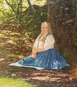 Frau mit langem blondem Haar in antiker blauer Hanftracht sitzt im Wald an einem Baum