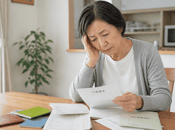 広島の60代女性が自宅のテーブルで戸籍や通帳、書類を見ながら少し困っている様子