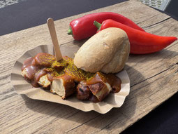 Currywurst mit Bröchen und Spitzpaprika im Hintergrund in braunem Pappschälchen auf Holzbrett