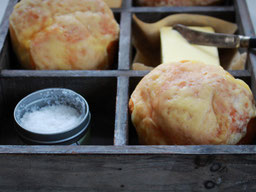 in einer Holzbox liegen 2 Käsebrötchen, ein Stück Butter, in dem ein Messer steckt, und ein Schälchen Salz