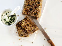 auf einer weißen Tischplatte liegt ein weißes Porzellanbrett, darauf ein Saatenbrot, 2 Scheiben sind abgeschnitten davor, rechts liegt ein Brotmesser mit Holzgriff, links steht ein weißes Porzellanschälchen mit Kräuterquark