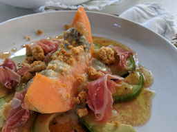 auf einem weißen Teller liegt eine gegrillte Melonenspalte mit käseCrumble und Serranoschinken