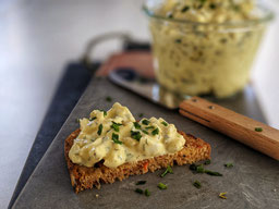 auf einem grauen Marmorbrett liegt eine Scheibe Vollkornbrot mit Eiersalat, im Hintergrund sieht man ein Messer mit Holzgriff und ein WeckglasEiersalat
