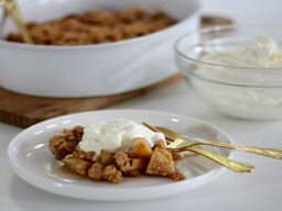 ein weißer Teller mit einer Portion ApfelCrumble mit JoghurtSahne, seitlich liegen eine kleine Gabel und ein kleiner Löffel, im Hintergrund eine weiße Schale