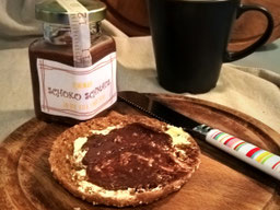 ein Toast mit Schokoladenaufstrich, auf einem Holzbrett liegend, daneben ein Messer mit buntem Griff, im Hintergrund eine Tasse und ein Glas Schokostreich