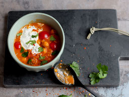 eine Schale Marrokanischer Linsentopf  mit Tomaten und gelben Paprika, einem Klecks Sauerrahm obenauf, die Schale steht auf einem grauen Marmorbrett, daneben liegt ein benutzter schwarzer Esslöffel
