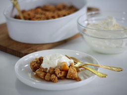 ein Dessertteller mit einem Stück Apfel Crumble mit Joghurtsahne, goldenes Besteck liegt daneben, im Hintergrund eine Auflaufschale und eine Glasschale mit Joghurtsahne