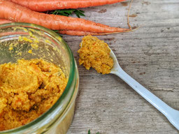 auf einem Holztisch steht ein Glas mit Karottenhummus mit Mandelmus, ein benutzter Löffel liegt rechts daneben, am oberen Rand sieht man 2 Karotten