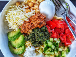 eine Bowl mit Nudeln, Kichererbsen, Gurken, Tomaten, geriebenen Käse und Pesto aus Karottengrün
