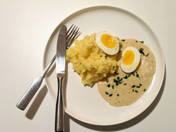 ein weißer Teller mit einem aufgeschnittenem Ei in Senfsauce mit Kartoffelstampf, Besteck liegt überkreuz rechts daneben