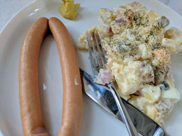 ein weißer Teller mit Kartoffelsalat und Heißwürstchen, Besteck liegt rechts überkreuz